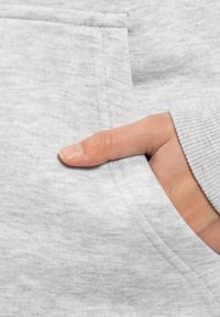 Jasnoszara bluza z kapturem z miękkiego, teksturowanego materiału. Ręka spoczywa w przedniej kieszeni, ukazując ściągacze i detale szwów.
