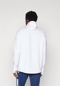 Wit met capuchon sweatshirt van gevoerde stof, ruime pasvorm en geribde manchetten, gedragen met blauwe sportbroeken met zijstrepen.