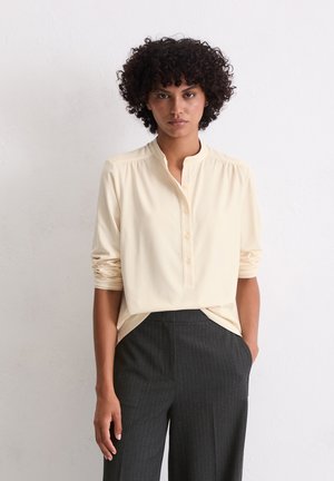 Crème blouse met lange mouwen en een mandarin col, knoopsluiting aan de voorkant en geplooide details op de schouders, gecombineerd met donkere broek met streepjes.