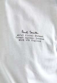 Hvid bomulds T-shirt med sort trykt tekst, inklusive "Paul Smith" og en adresse i London. Glat tekstur med minimal detailing.