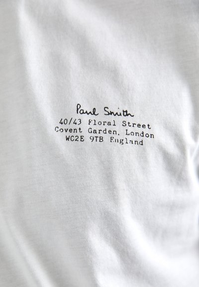 Fehér pamut póló fekete nyomott felirattal, amely tartalmazza a "Paul Smith" nevet és egy londoni címet. Simává textúrált, minimális díszítéssel.