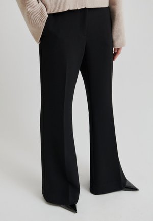 Inimene, kes kannab musti laia säärega pükse ja musti teravate otstega kingi, kombineeritud beežika soonikkoes pika varrukaga kampsuniga.