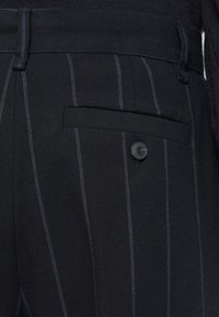 Pantaloni a righe nere con strisce verticali blu, una tasca a filetto e un dettaglio con bottone nero di lato. Realizzati in tessuto testurizzato.