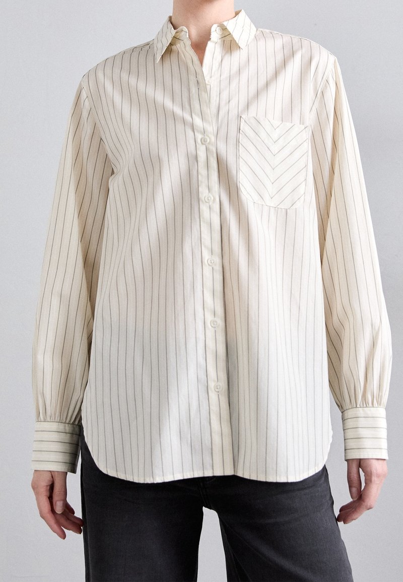 Personne portant une chemise crème à manches longues avec de fines rayures verticales et une poche poitrine à motif chevrons.