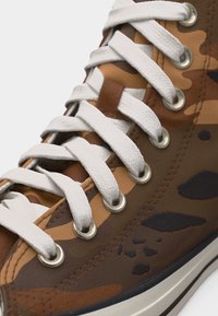 Visoka tenisica s uzorkom smeđe i bež kamuflaže, bijelim vezicama provučenim kroz metalne rupice i bijelim gumiranim potplatom s crnom prugom.