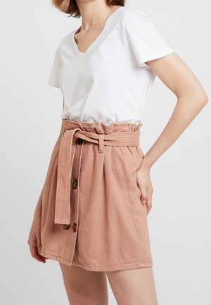 Jupe en coton rose pâle avec taille nouée, dotée d'une patte de boutonnage sur le devant et de plis décoratifs. Associée à un t-shirt blanc à col en V.