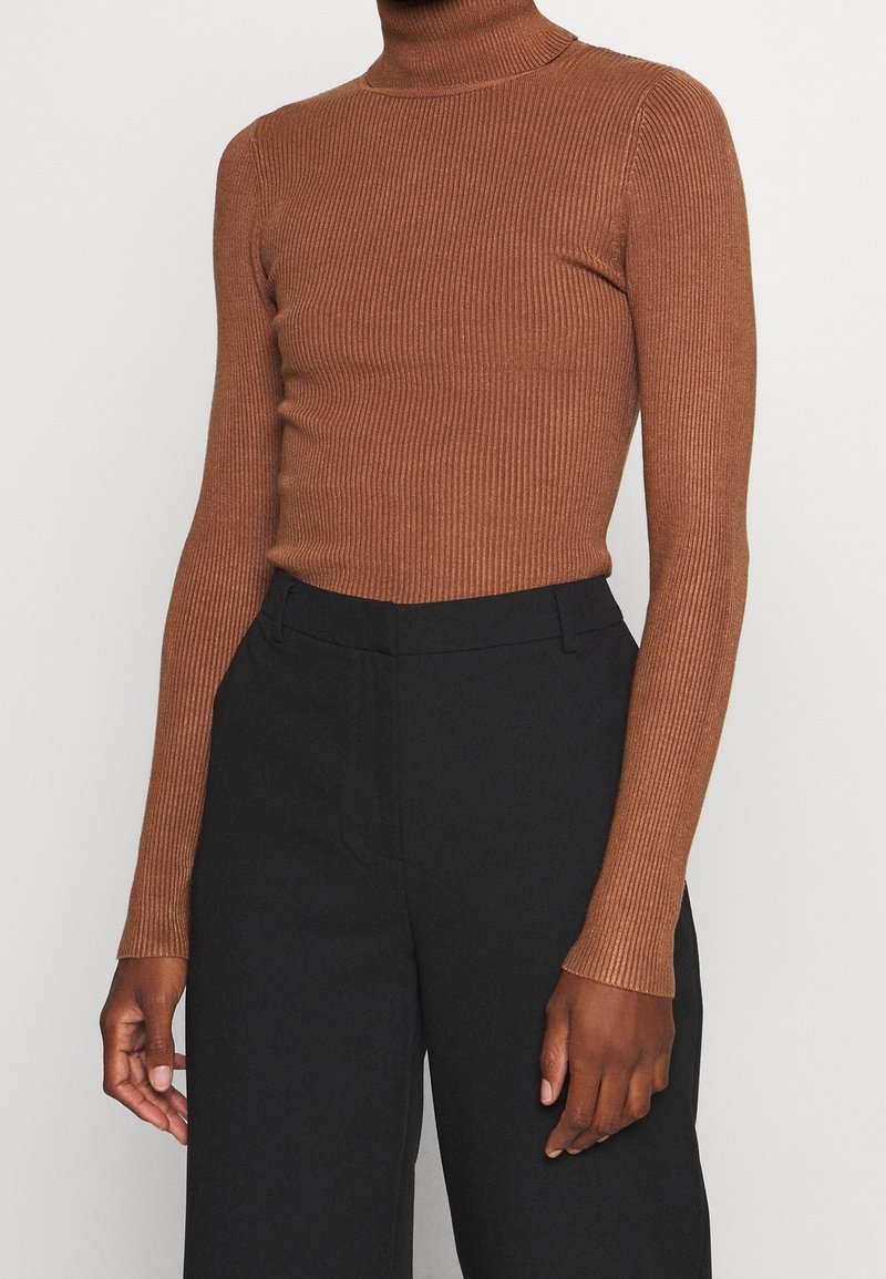 Pull à col roulé côtelé marron porté avec un pantalon noir large taille haute sur une personne devant un fond uni.