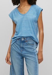 Modrý melírovaný bezrukávový top s kulatým výstřihem, spárovaný se světlými modrými volnými džínami s přední kapsou a zapínáním na knoflík.