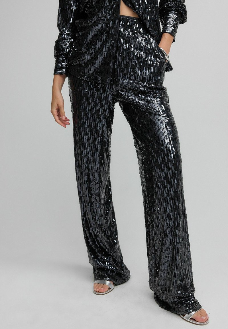 Femme portant un pantalon noir à sequins et une veste assortie, avec des sandales argentées à bouts ouverts, debout, une main dans la poche.