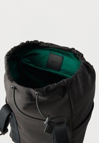 Sac à cordon en tissu noir avec une doublure intérieure verte et une petite étiquette rectangulaire cousue à l'intérieur, près du bord supérieur.