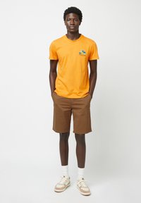 T-shirt arancione a maniche corte con una grafica blu e bianca, abbinato a pantaloni corti marroni fino al ginocchio. Il modello indossa calze bianche e scarpe da ginnastica beige.