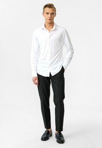 Witte lange mouwen hem met een button-down kraag, gecombineerd met slim-fit zwarte broek en zwarte instappers, tegen een effen achtergrond.