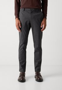 Hombre vestido con pantalones oscuros de corte ajustado, mocasines marrones y una camisa de manga larga marrón, de pie contra un fondo claro y liso.