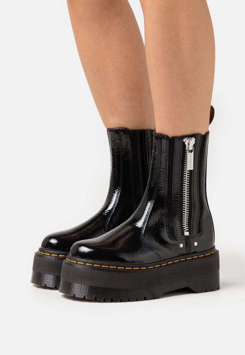 Bottines à plateforme en cuir verni noir brillant avec semelles épaisses à rainures, fermeture éclair latérale et coutures jaunes portées sur jambes nues.