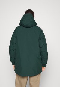 Persoon met de rug naar voren, gekleed in een donkergroene gewatteerde jas met capuchon en beige broek, tegen een effen lichte achtergrond.