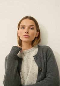 Cárdigan gris con puños acanalados, que presenta tres botones y una textura suave, superpuesto a un suéter blanco moteado. Cuello y diseño sencillos.