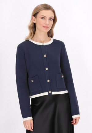 Navy blue knitted cardigan with cream accents, round neckline, five gold buttons, and two front pockets. Cropped design with long sleeves.