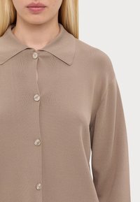 Beżowa koszula zapinana na guziki z dużym kołnierzem, wykonana z teksturowanej tkaniny, z zapięciem na guziki z przodu i długimi rękawami.