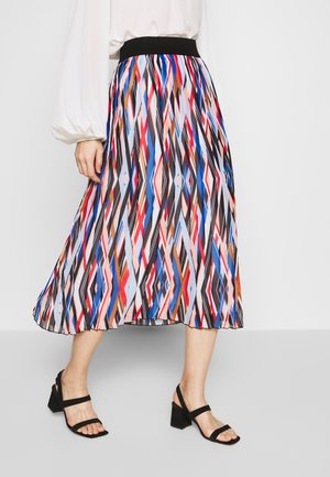 Femme portant une jupe midi plissée à motifs géométriques colorés avec une ceinture noire, un chemisier blanc et des sandales à talons noires en marchant.