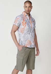 Camicetta a maniche corte grigia chiara con motivi di foglie arancioni e grigi; abbinata a pantaloni cargo verde oliva con tasche laterali.