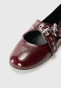 Scarpa in pelle patinata bordeaux con punta arrotondata, ampia fascia con occhielli in argento e fibbia, e un design a taglio aperto.