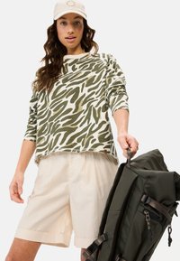 Grøn og creme dyreprint langærmet skjorte med frynset kant, parret med lysebeige shorts og en mørkegrøn rygsæk.