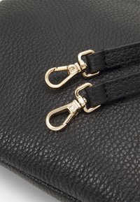 Bolso de cuero negro con textura que presenta dos clips metálicos dorados sujetos a las correas. Destaca por su grano visible y detalles de herrajes lisos.