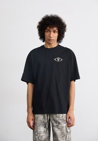 Black cotton T-shirt with a loose fit and short sleeves, featuring a small intricate design on the left chest. Textured gray camo cargo pants.