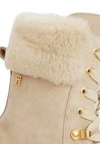 Botín de ante beige con un suave dobladillo forrado de pelo, herrajes dorados y cordones beige. Acentuado con un pequeño detalle de logo en el costado.