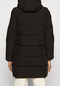 Person, der eine lange schwarze gefütterte Jacke mit Kapuze und horizontalen Nähten trägt, steht mit dem Rücken zur Kamera, mit weißen Hosen, die darunter sichtbar sind.