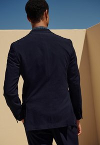 Navyfarbener, strukturierten Blazer von hinten gezeigt, mit einem Schlitz, taillierter Passform und sichtbaren Kontrastärmel darunter.