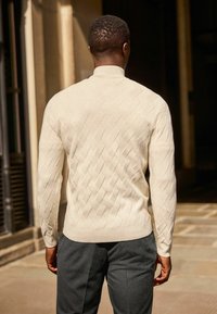 Suéter de punto crema claro con un patrón de diamante texturizado, que presenta un cuello y puños acanalados, combinado con pantalones gris oscuro.