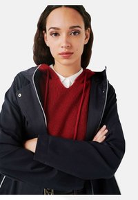 Giacca impermeabile nera con zip argentata, sovrapposta a un maglione di lana rosso con colletto bianco. Il modello ha le braccia incrociate.