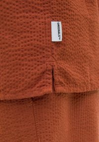 Roodbruine gestructureerde stof met een ruitpatroon, met een klein label dat aan de rand is genaaid en "ORIGINALS™" leest. Het op maat gemaakte zoomdetail is zichtbaar.