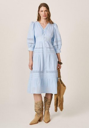 Mujer con vestido midi azul claro de mangas abullonadas, detalles con botones, sosteniendo una bolsa marrón con flecos y con botas de vaquero marrones con recortes florales.