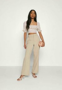 Top crop floral claro com mangas bufantes, combinado com calças de cintura alta listradas em bege e sapatos de salto brancos com tiras, segurando uma bolsa rosa.