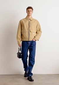 Beige zip-up jakke med to frontlommer, kombinert med mørkeblå jeans. Modellen holder en liten svart veske og har på seg svarte sko.