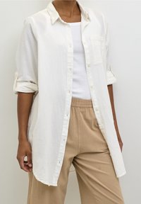 Chemise blanche légère à boutons, avec une poche poitrine et des manches retroussées, portée avec un pantalon beige à taille élastique, offrant une coupe décontractée.