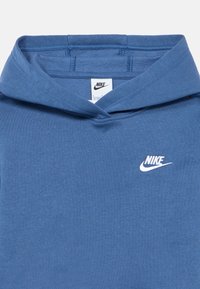 Sininen pehmeästä kankaasta valmistettu huppari, jossa on kenguruntasku, kiristysnyörillä varustettu huppu ja valkoinen Nike-logo vasemmassa rintataskussa.