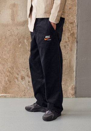 Person iført sorte Nike Sportswear-bukser med hånden i lommen og grå sneakers, stående op ad en tekstureret beige væg.