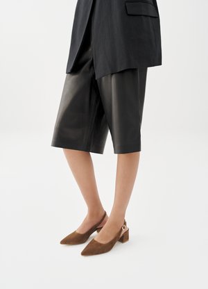 Nő fekete, térdig érő bőr rövidnadrágban, fekete blézerben és barna, alacsony blokktalpú, szarvasbőr szandálcipőben.