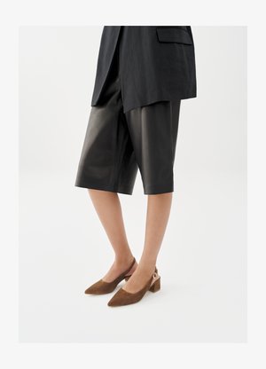 Kobieta w czarnych, skórzanych szortach do kolan, czarnym marynarce oraz brązowych zamszowych sandałach na niskim obcasie na pasku z tyłu.