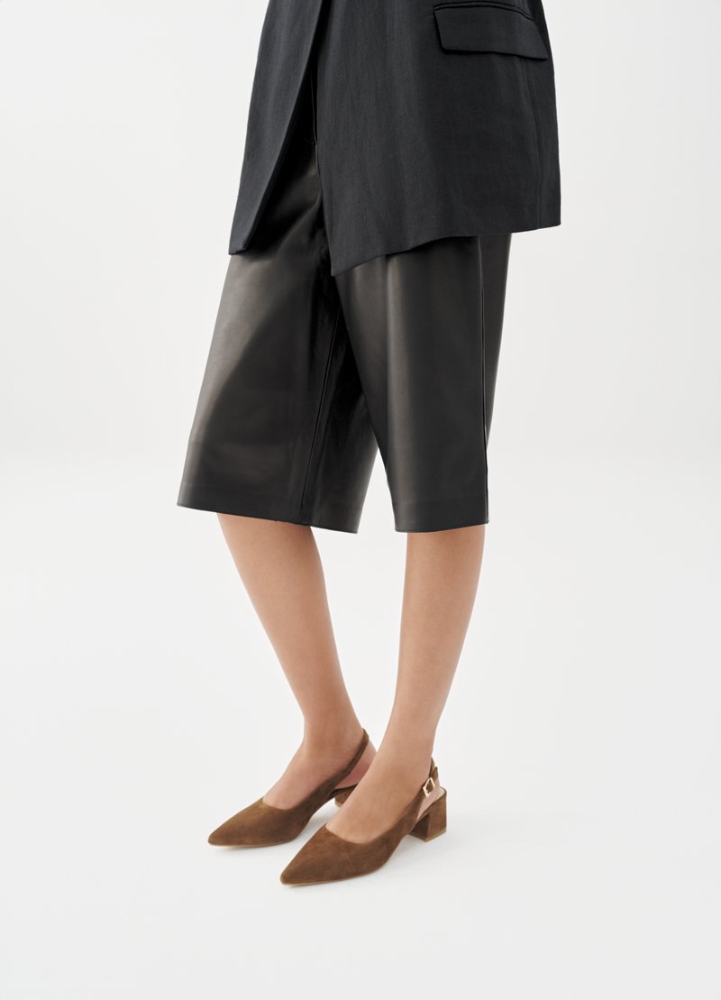 Frau trägt schwarze knieumspielende Lederhosen, ein schwarzes Sakko und braune Wildleder-Slingback-Schuhe mit niedrigem Blockabsatz.