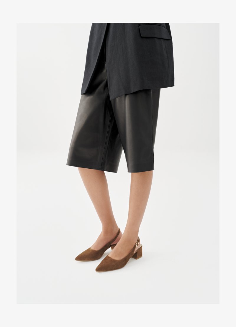 Frau trägt schwarze knieumspielende Lederhosen, ein schwarzes Sakko und braune Wildleder-Slingback-Schuhe mit niedrigem Blockabsatz.