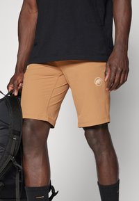 Beige atletiska shorts med en figurnära design, slät yta och logotypacc jac på låret. Bärs med en svart t-shirt och håller en ryggsäck.
