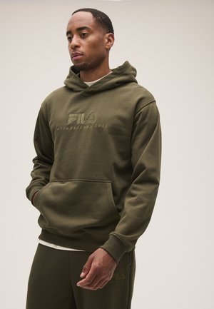 Hombre con una sudadera con capucha FILA verde oliva y pantalones a juego, de pie con las manos en el bolsillo de la sudadera contra un fondo liso y claro.