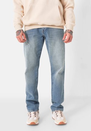 Hellblaue Jeans aus Denim mit lockerer Passform, ausgestattet mit dezenten Gebrauchsspuren und einem klassischen Fünf-Taschen-Design. Kombiniert mit neutral gefärbten Sneakers.