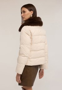 Giacca imbottita crema con colletto in pelliccia marrone, caratterizzata da cuciture verticali, due tasche laterali e vestibilità comoda. Indossata con una gonna marrone.