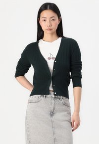 Dark green ribbed cardigan with a V-neck, long sleeves, and button closure. Worn over a white T-shirt with cherry graphic and paired with a grey skirt.