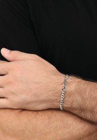 Pulsera de cadena de metal plateado con eslabones entrelazados, superficie lisa, llevada en una muñeca con vello claro, contra un fondo de camisa negra.